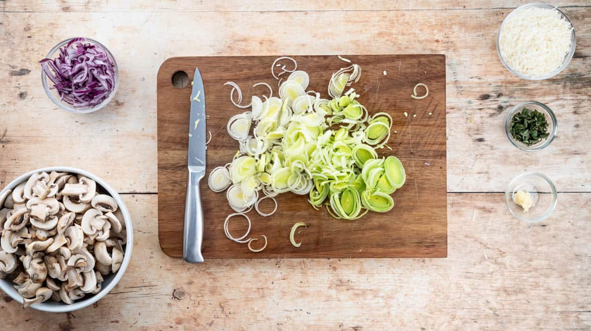 A wooden chopping board with sliced leeks and a sharp knife sat upon it. Around the board are sliced mushrooms, sliced onions, chopped sage, crushed garlic, and grated parmesan.