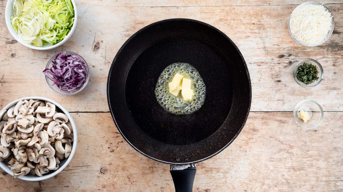 A frying pan with butter that is melting inside. Surrounding the pan are some of the ingredients needed to make the creamy mushroom sauce.
