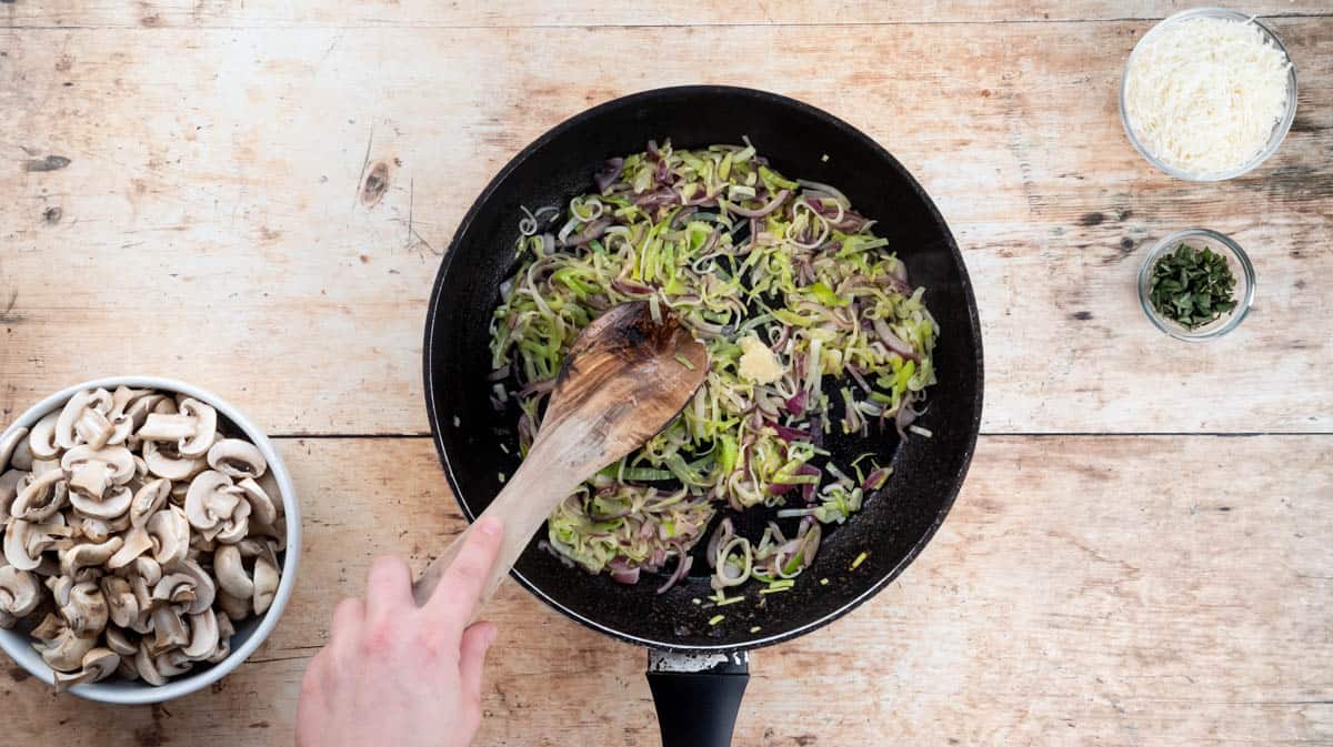 A hand is using a wooden spoon to stir the leeks and onions as they saute in a pan.