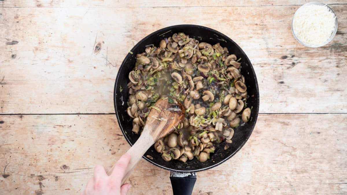 A hand is using a wooden spoon to stir wine into the mushroom sauce mixture.