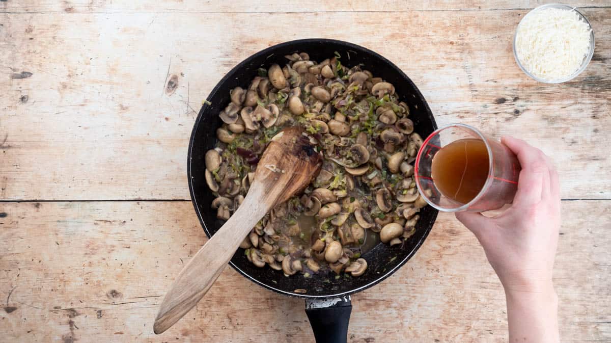 A hand is tipping chicken stock into a frying pan containing cooked mushrooms, leeks and onions for the mushroom sauce.