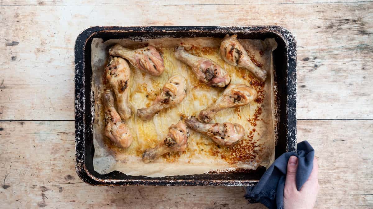 A hand is using a blue cloth to hold a hot tray of baked chicken drumsticks.