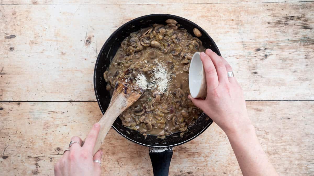 A hand is tipping parmesan cheese into the frying pan that contains other ingredients for the creamy sauce.