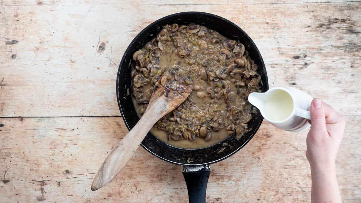 A hand is tipping a white jug of double cream into the mushroom sauce in a black frying pan.