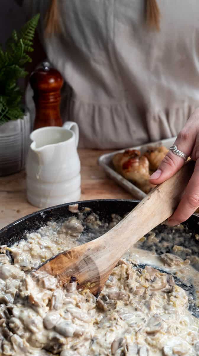A person is using a wooden spoon to stir the creamy mushroom sauce inside a frying pan.