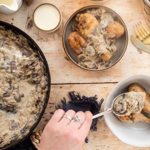 A hand is spooning mushroom sauce on top of chicken drumsticks in a white bowl.