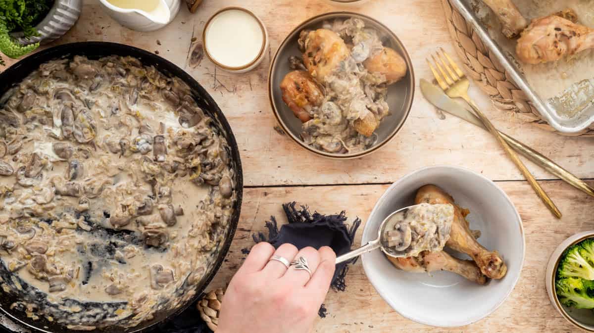 A hand is spooning mushroom sauce on top of drumsticks in a bowl. There are much creamy chicken drumsticks in another bowl, the mushroom sauce in a frying pan, and more chicken drumsticks on a baking tray.