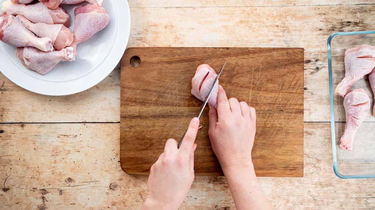 A hand is using a sharp knife to score diagonal cuts across each drumstick on a wooden chopping board.