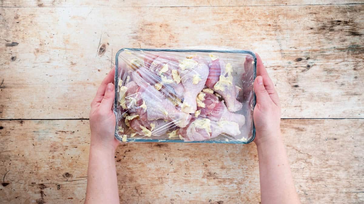 Two hands are holding a rectangular glass dish filled with chicken drumsticks that have been covered in a sage butter mixture. The dish is covered with cling film.