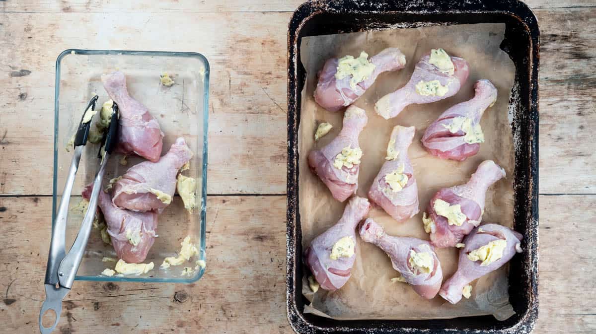 An overhead view of chicken drumsticks covered in butter and laid in a single layer inside a baking tin that has been lined with baking paper. Next to the tin is a glass bowl with tongs and more chicken drumsticks inside.