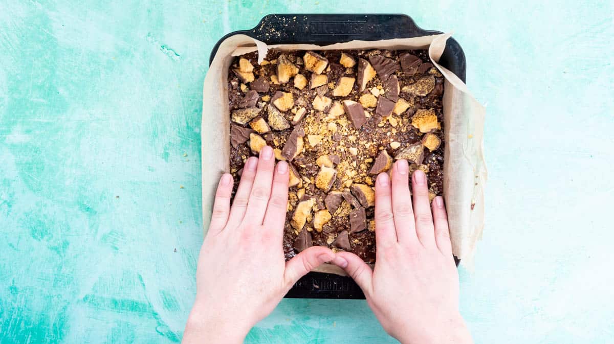 Two hands our pressing broken Crunchie pieces into the top of the Tiffin mixture inside a baking tin.