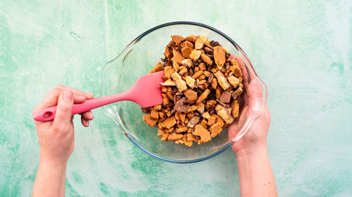 A hand is using a pink spatula to mix the dry ingredients of the Tiffin together inside a glass bowl.