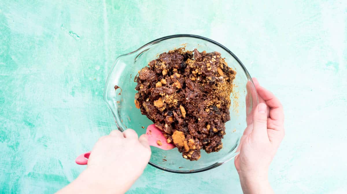 A hand is using a pink spatula to mix the Tiffin mixture together inside a glass bowl.