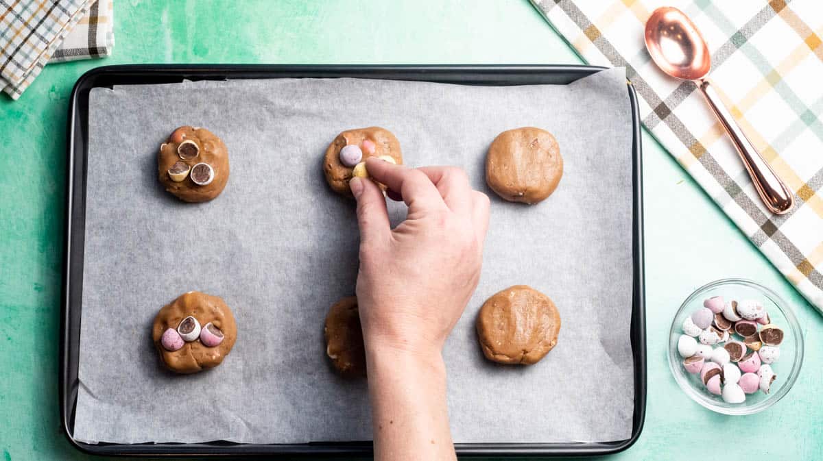A hand is adding chopped mini eggs to the top of flattened cookie dough balls.