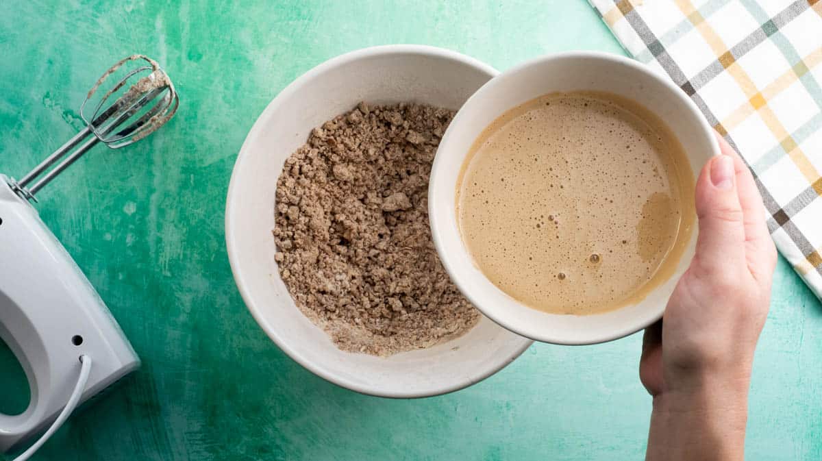 A hand is tipping creamed sugar and egg in with the other cookie ingredients in a white bowl.