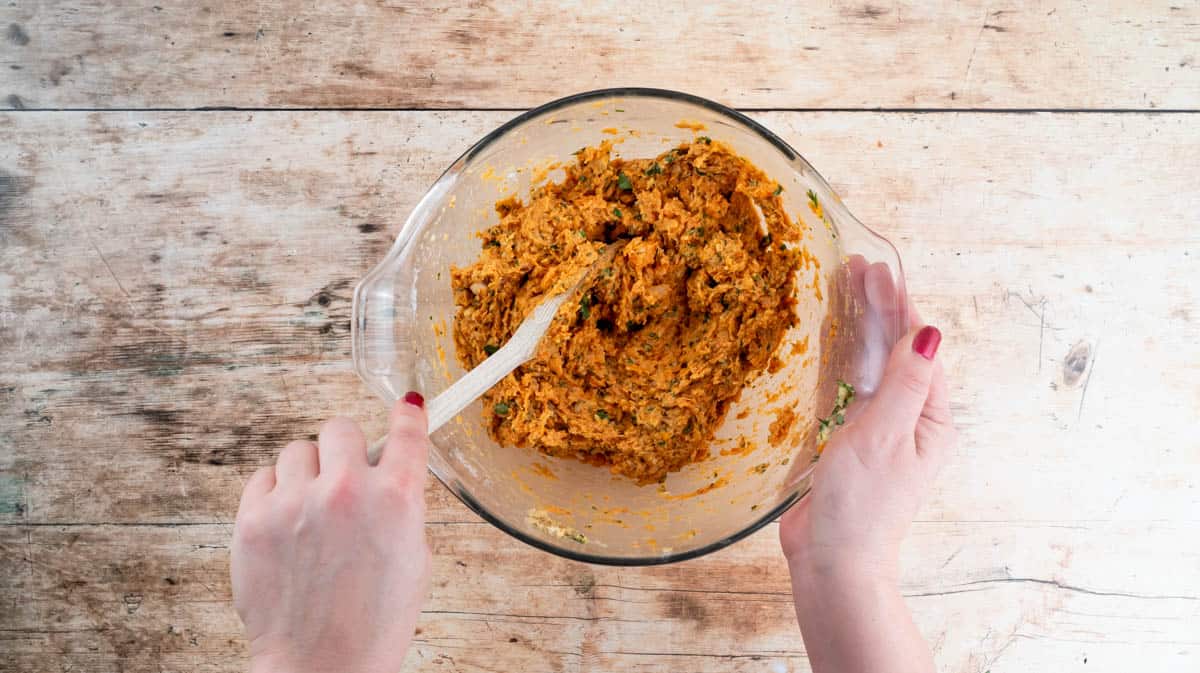 A hand is using a wooden spoon to mix the mashed falafel ingredients together in a glass bowl.