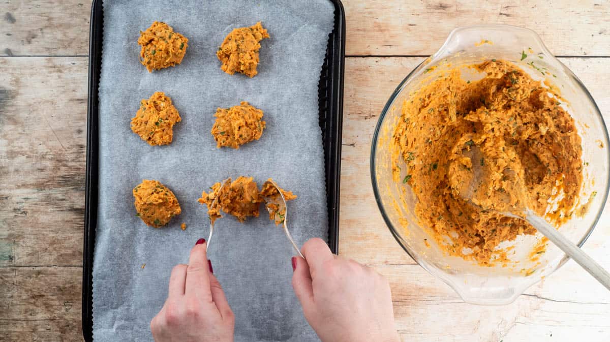 Two hands are using spoons to ball the falafel mixture onto a lined baking tray.