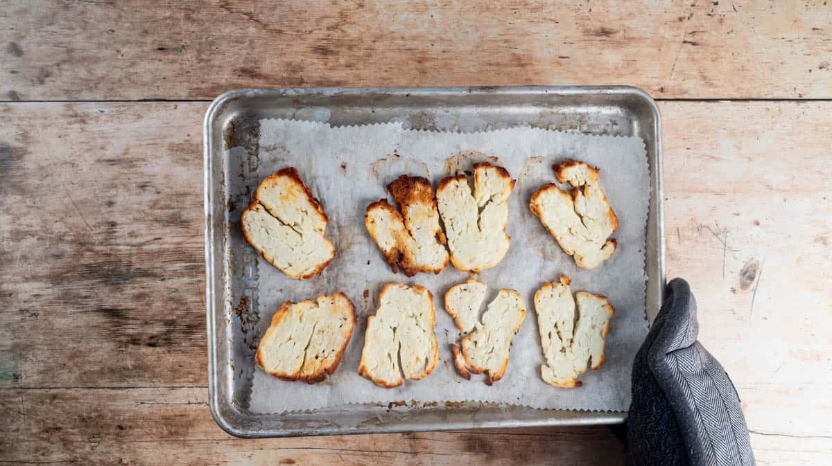 A gloved hand is holding a baking tray with baked halloumi slices on it.