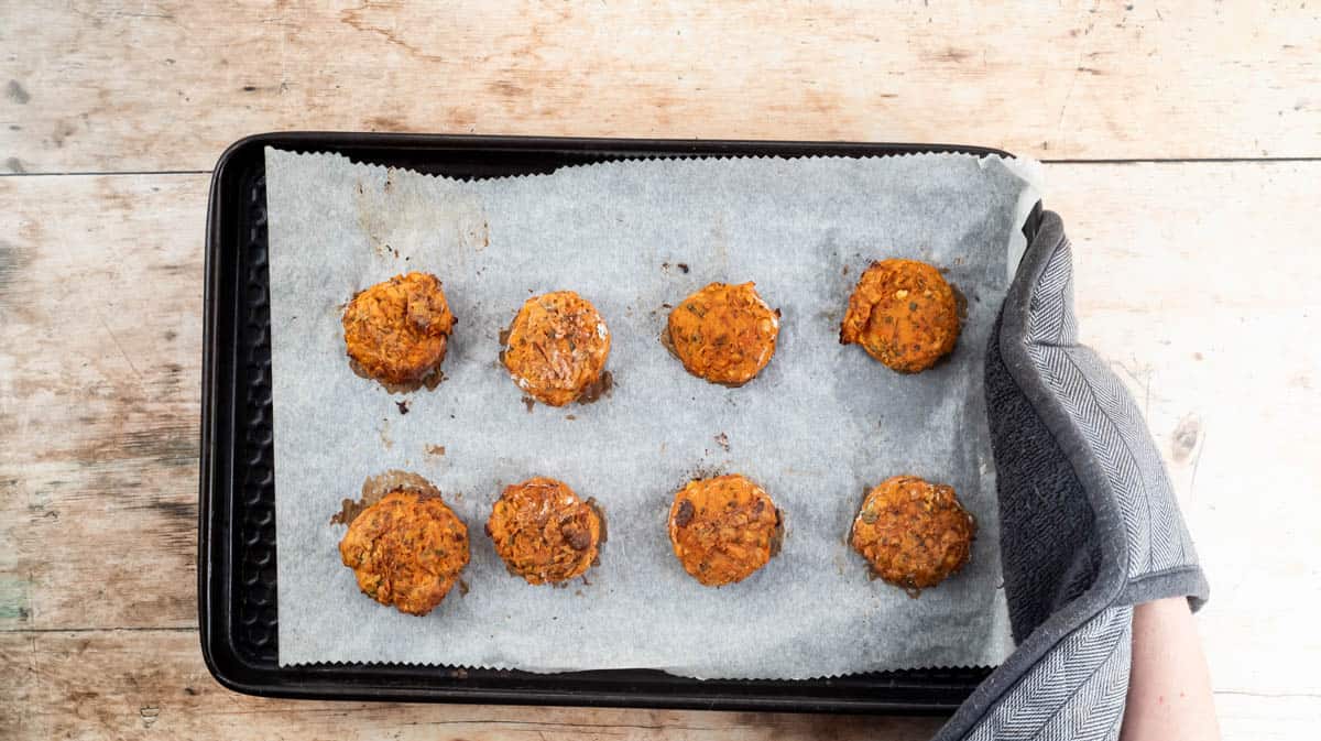 A gloved hand is holding a baking tray with baked falafels on it.