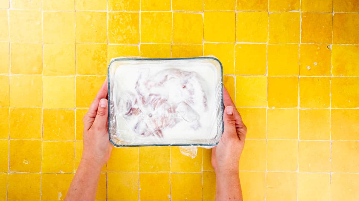 Two hands are holding a glass dish with buttermilk marinated chicken inside.