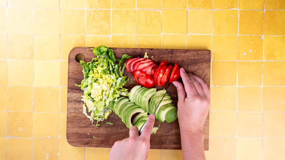 A hand is using a sharp knife to slice tomato, lettuce and avocado on a wooden board.