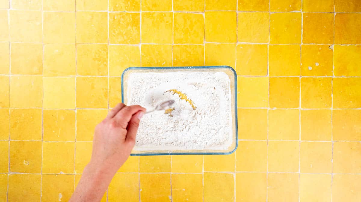 A hand is using a spoon to mix the flour coating ingredients for the chicken together in a glass dish.