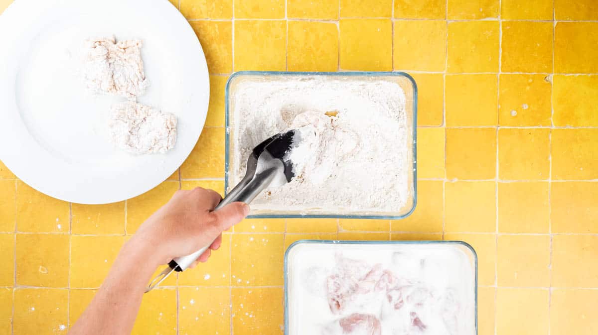 A hand is using a pair of tongs to place the chicken in the flour mixture to coat it before frying.