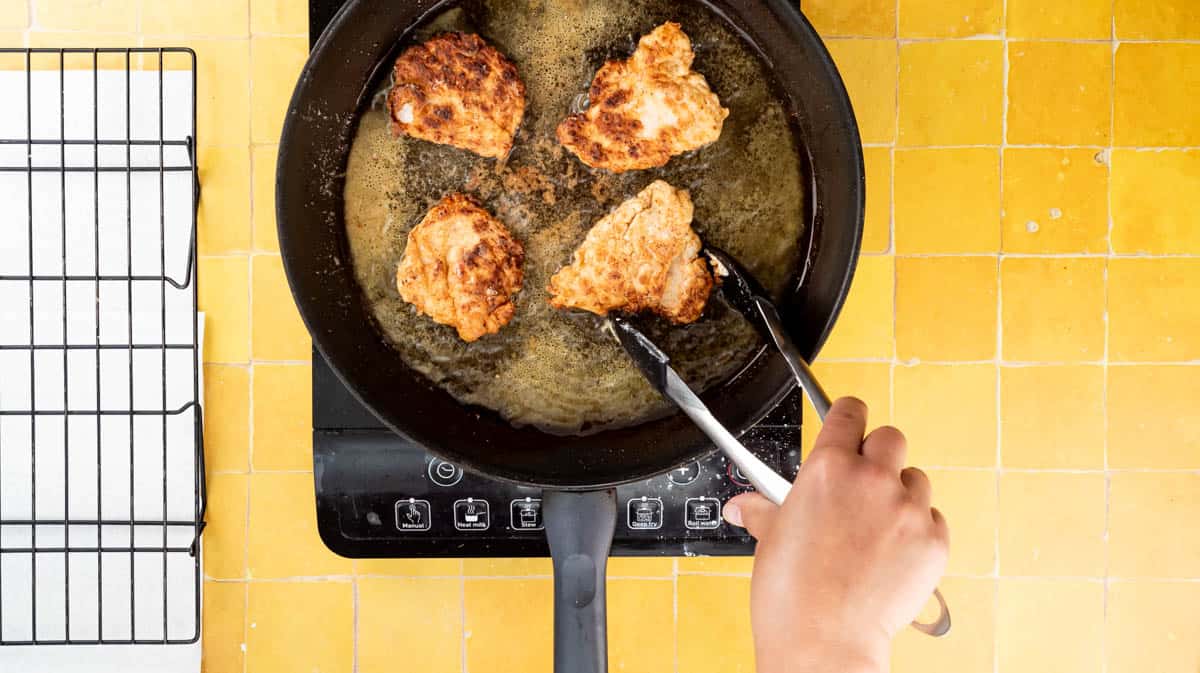 A hand is using a pair of tongs to turn the chicken in the oil in a frying pan as it cooks.