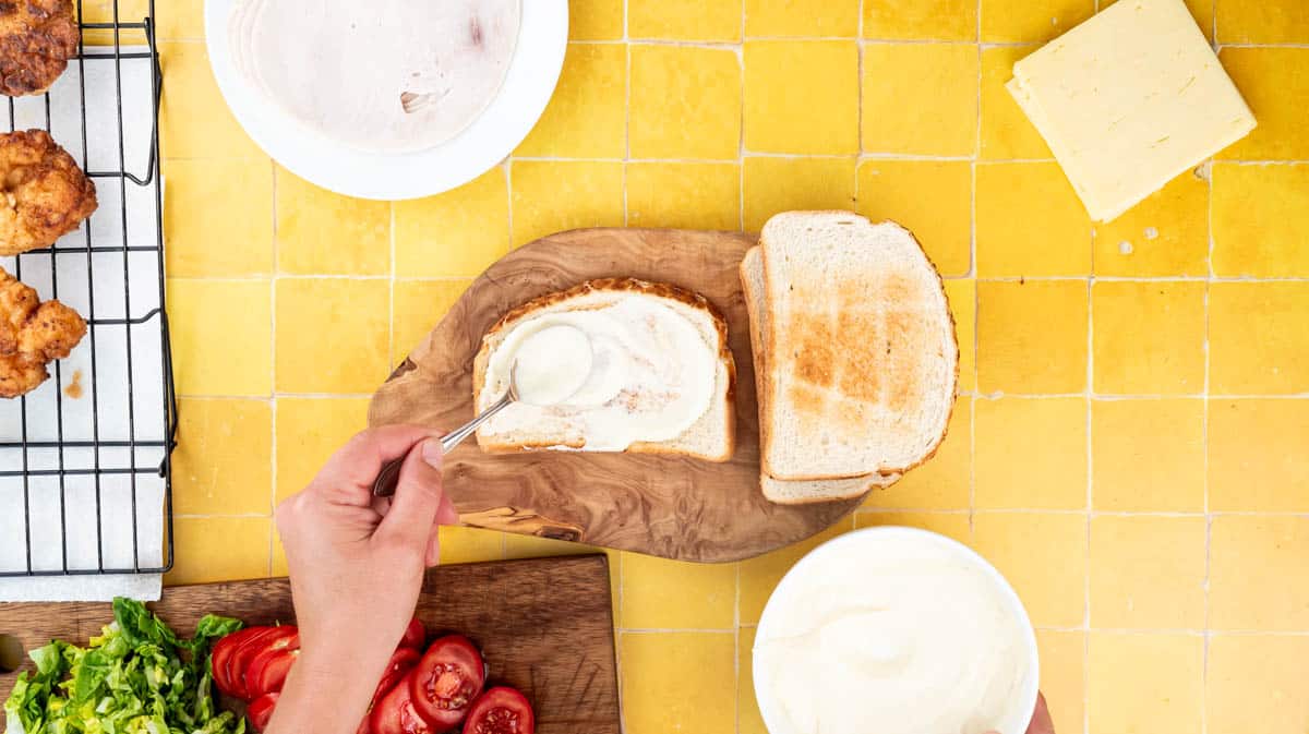 A hand is using a spoon to spread a toasted slice off bread with mustard Mayo.