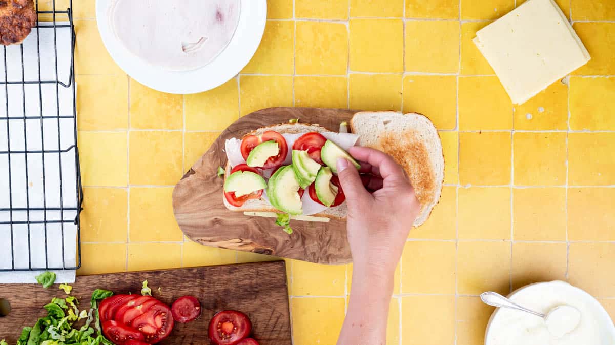 A hand is placing sliced avocado in the club sandwich.