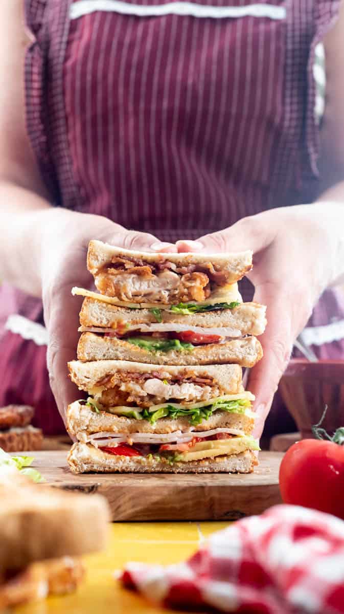 A person is holding a stacked fried chicken club sandwich on a wooden board to show the layers.