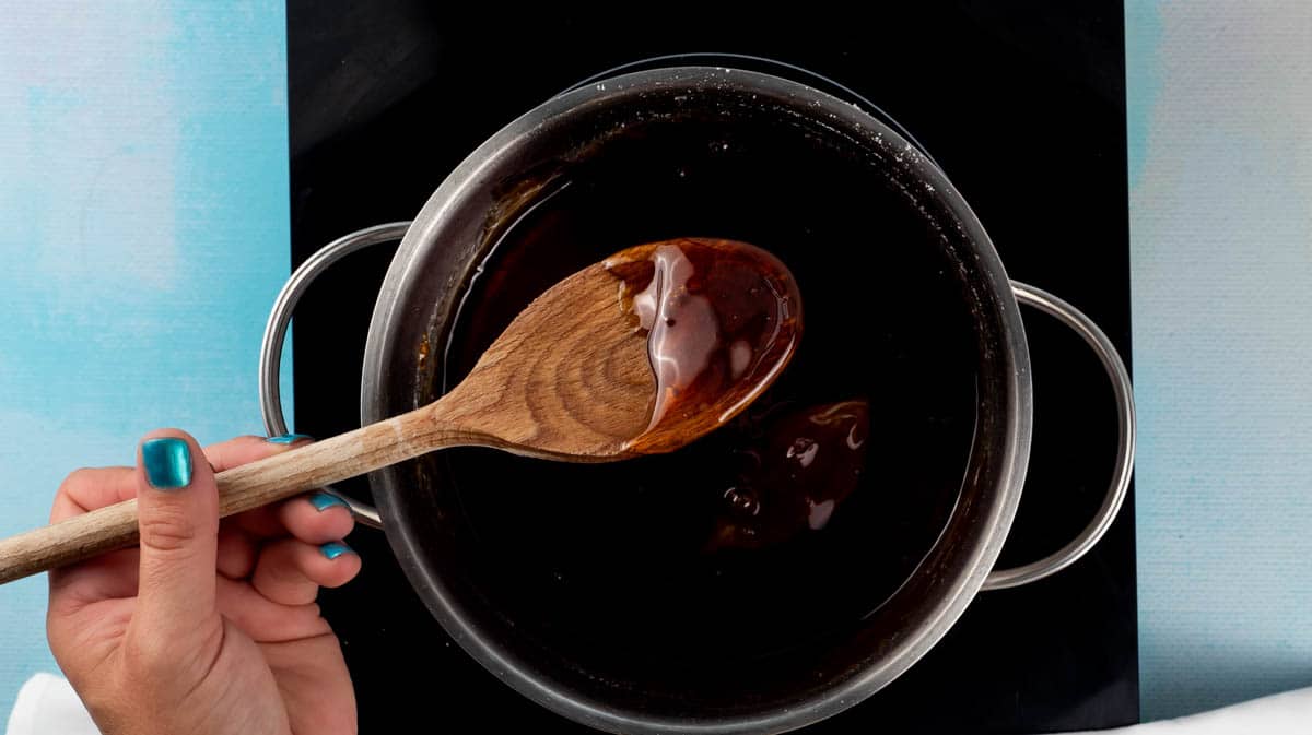 A hand lifting the wooden spoon from the golden syrup to show the thick consistency.