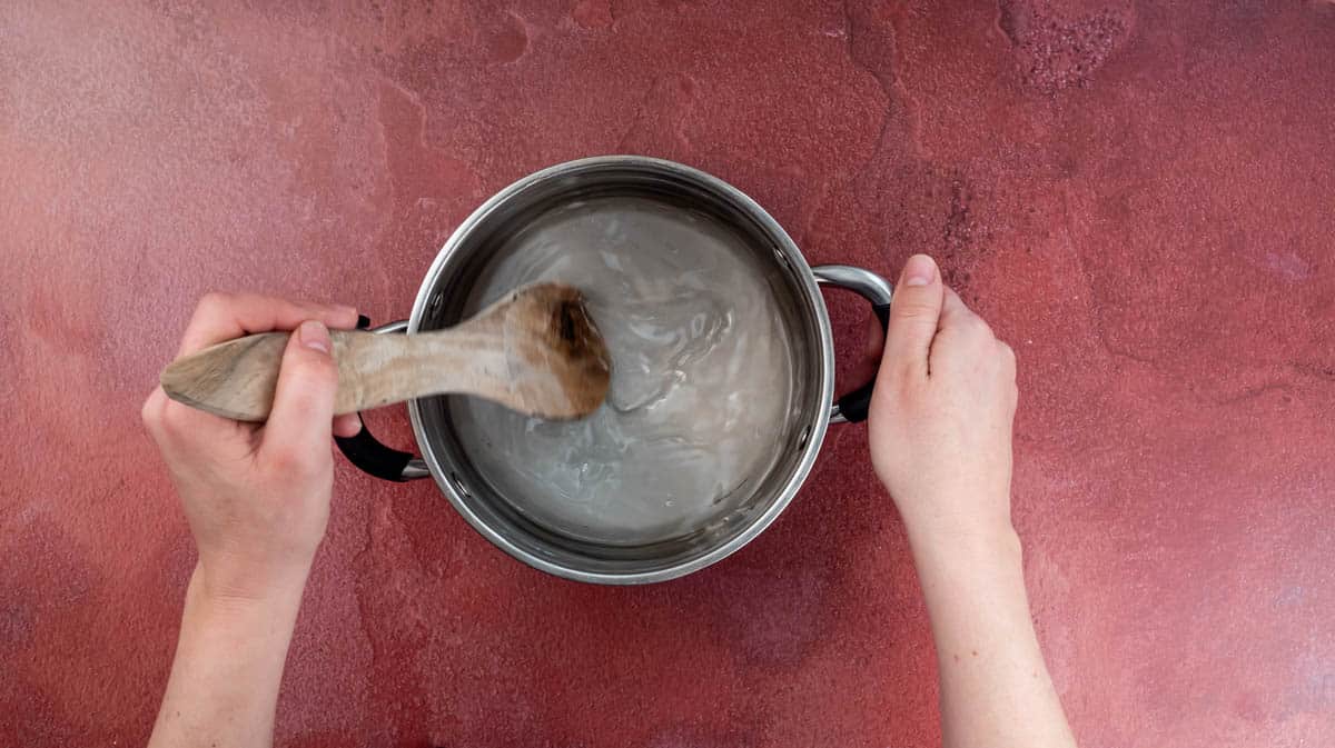 A hand is using a wooden spoon to stir sugar and water together in a saucepan.