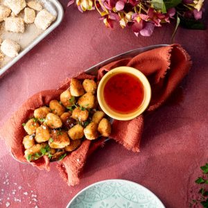 A bowl of halloumi nuggets with a sweet chilli sauce on the side of the pile. There are uncooked nuggets and decorative items also in the photo.