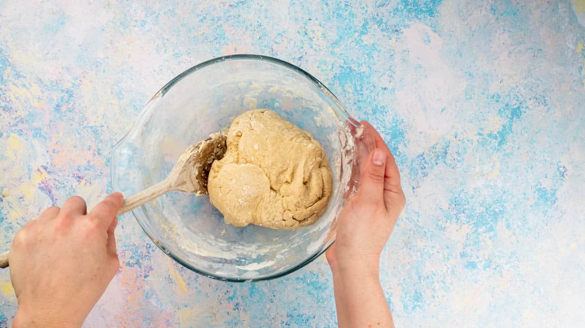 A hand is using a wooden spoon to mix the flour and wet ingredients together to form a dough.