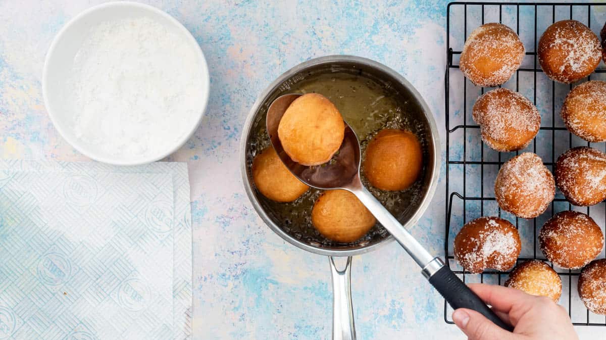 A hand is using a slotted spoon to lift a fried doughnut from a saucepan filled with oil.
