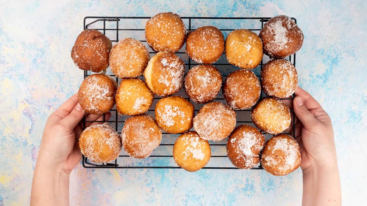 Two hands are holding the sides of a wire rack that's filled with sugar-coated doughnuts.