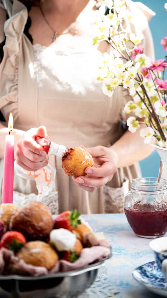 A person is using a piping bag to fill a doughnut with jam. In the front of the picture there are decorative items.