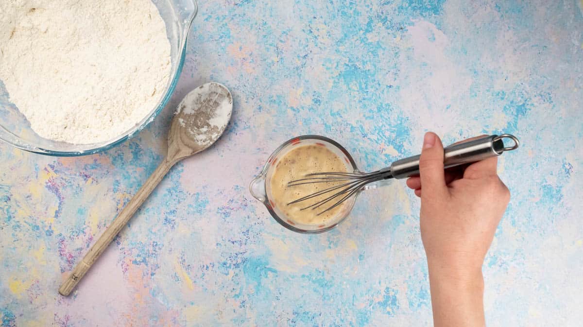 A hand is using a whisk to mix together the milk-yeast mixture with an egg.