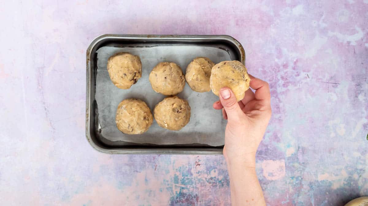 A hand is placing Kinder cookie dough balls into a lined baking tray for freezing.