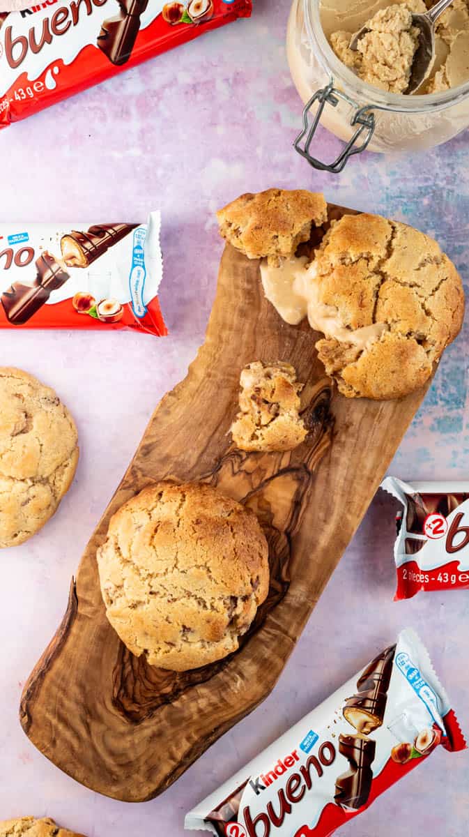 A wooden board with a broken open Kinder Bueno cookie to show the centre. There are other whole cookies and Kinder Bueno chocolate bars around the cookies.