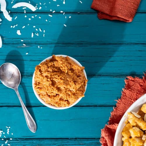 A pot of korma Curry paste next to a spoon, in the corner there is a bowl of Curry and ingredients surrounding the photo.