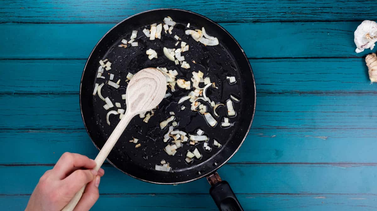 A person is using a wooden spoon to stir onion in a frying pan as it sautees.