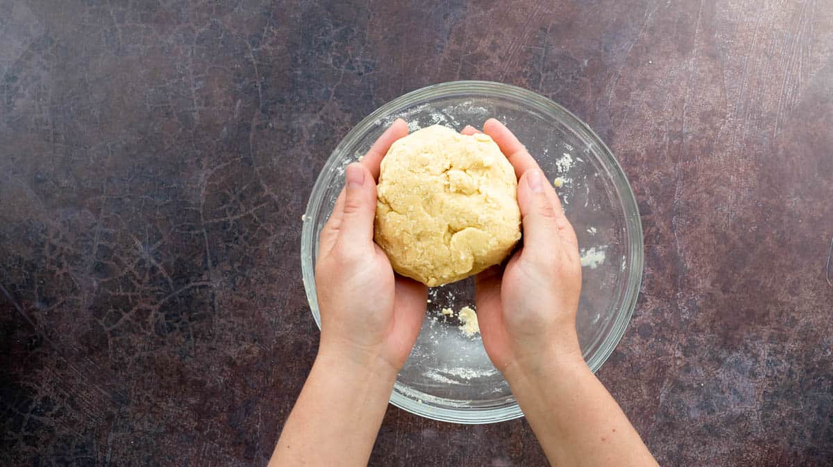Two hands are cupping a ball of shortcrust pastry dough.