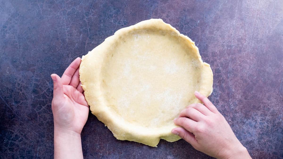 Two hands are pressing the rolled out shortcrust pastry into a round quiche tin.