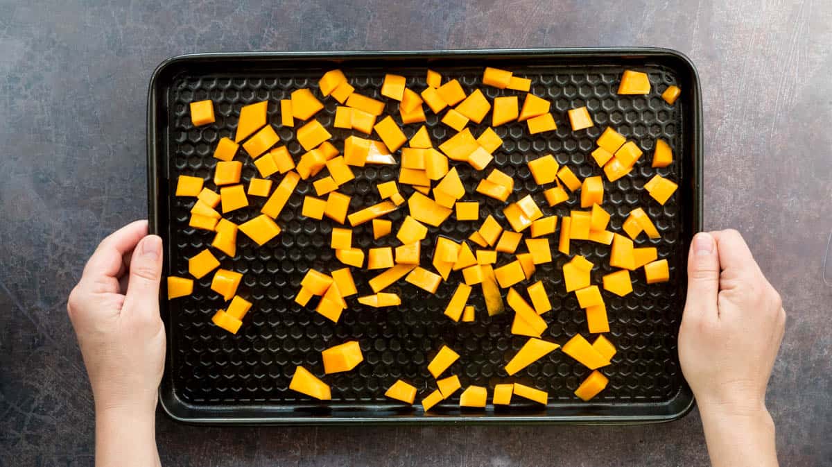 Two hands are holding a baking tray covered in cubes of butternut squash ready to take to the oven.