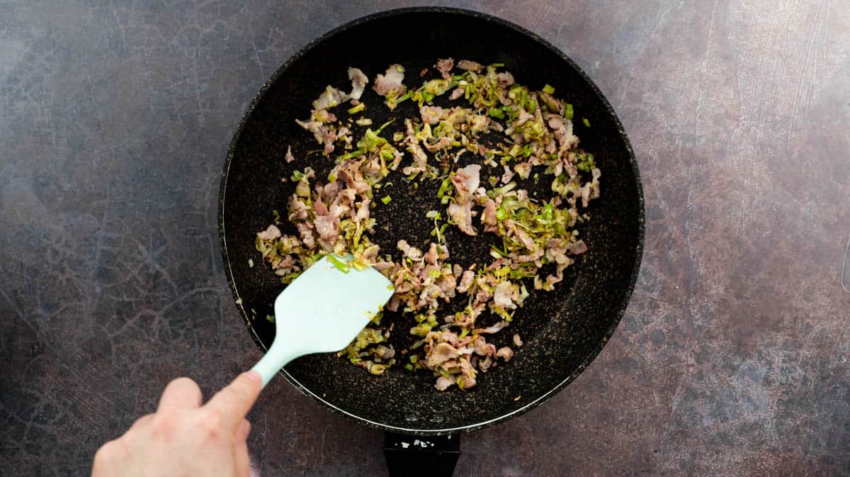 A hand is using a blue spatula to stir the leek and bacon in a frying pan as it cooks.