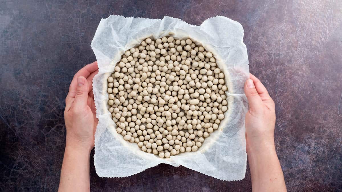 Two hands are holding the pastry base that has been filled with baking beans and lined with baking paper.