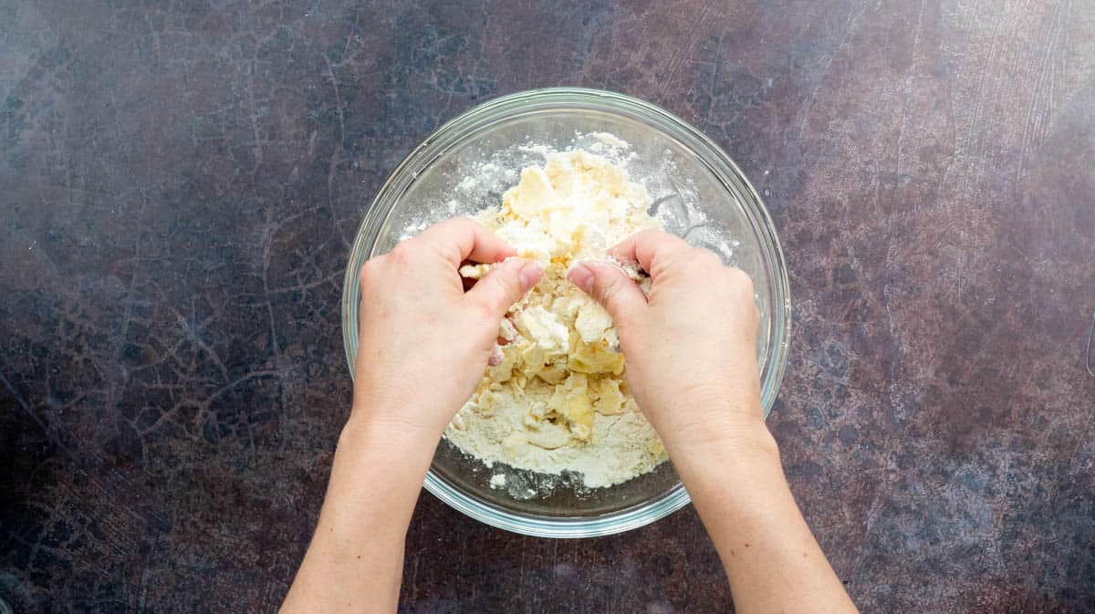 Two hands are rubbing butter into the flour in a glass bowl.
