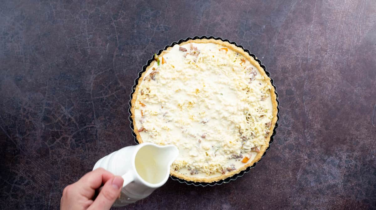 A hand is pouring the egg-cream mixture into the quiche case that has been filled with the other ingredients.